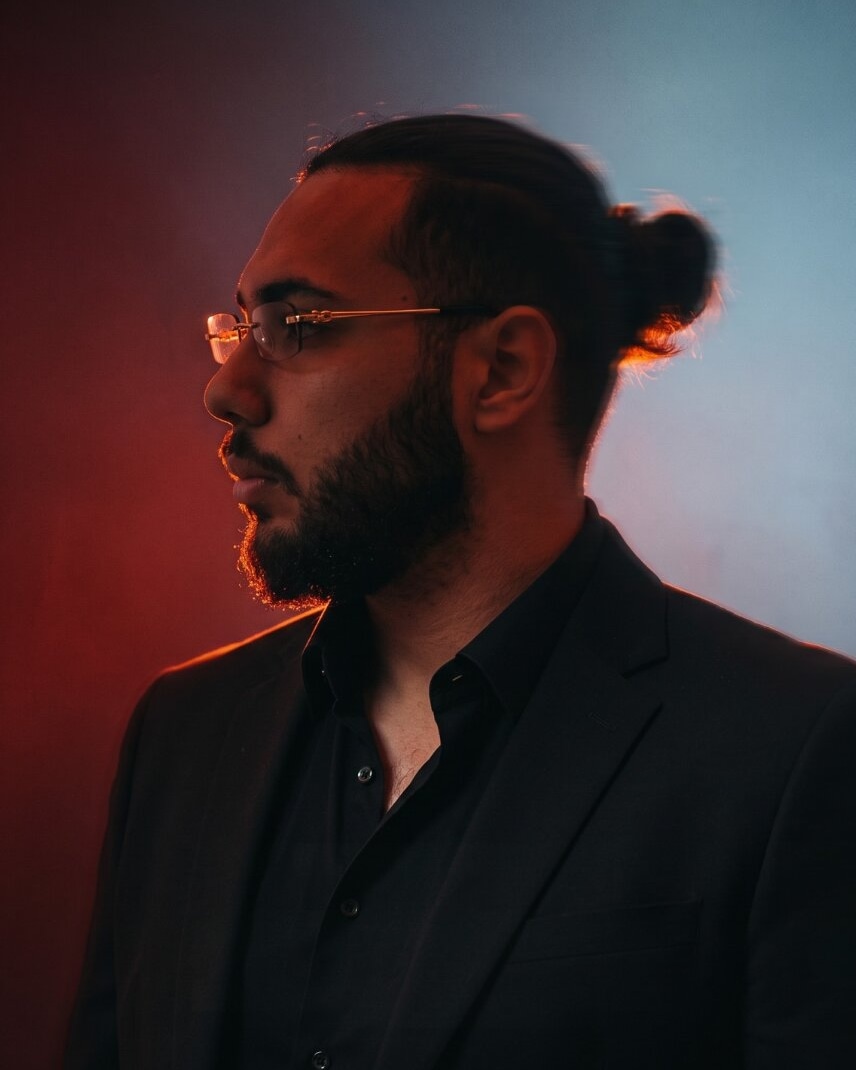 Yousef Abdo in profile, wearing glasses and a black suit, with red and blue lighting.